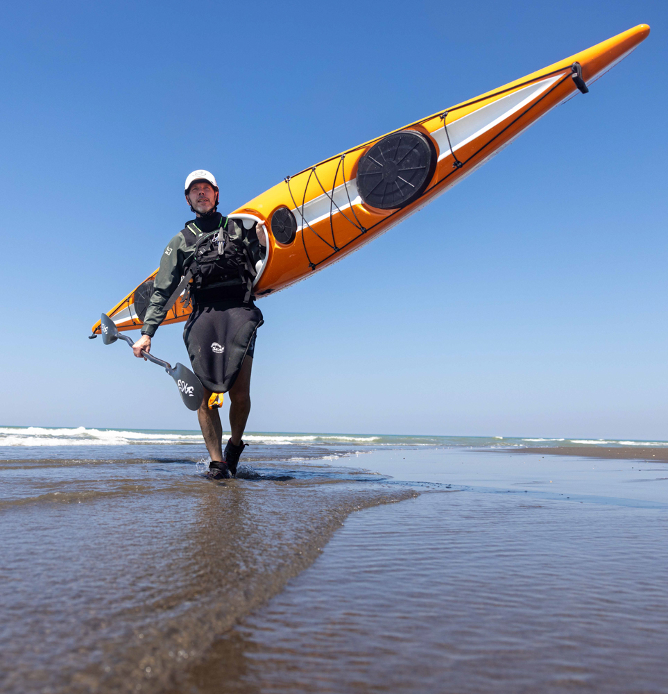 A paddler carrying an Edge Alnes down the beach